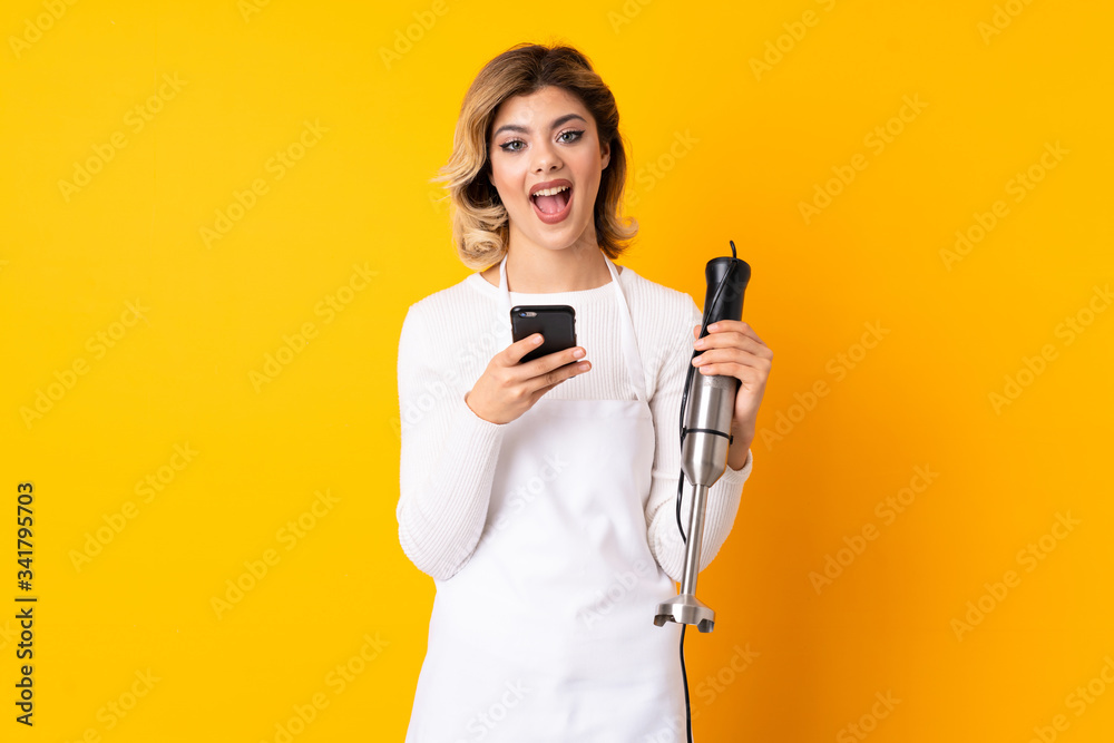 Girl using hand blender isolated on yellow background surprised and sending a message Girl using hand blender isolated on yellow background surprised and sending a message