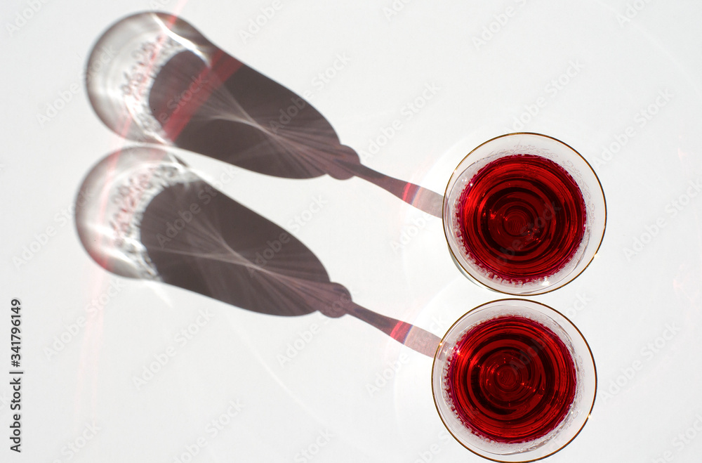 Top view. Flat lay. Two crystal glasses with red wine. Glass goblets with alcohol against background of white wall. White linen tablecloth. Direct sunlight, long shadows. Selective focus image. Top view. Flat lay. Two crystal glasses with red wine. Glass goblets with alcohol against background of white wall. White linen tablecloth. Direct sunlight, long shadows. Selective focus image.