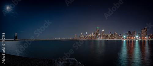 A beautiful panoramic shot of the Chicago skyline and city at night from the curved pier at North Avenue beach with light radiating from the moon and lights reflecting on the water of Lake Michigan.