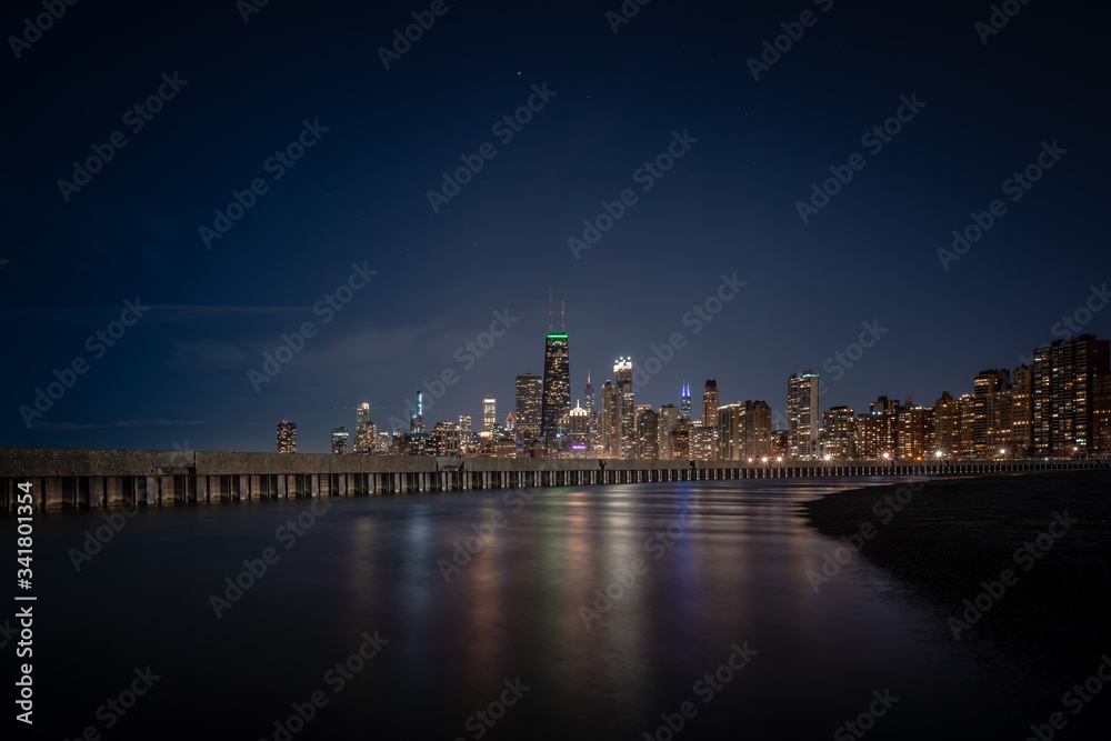 Colorful building lights reflecting off of standing water adjacent to corrugated steel sheet piling and concrete pier with the Chicago Skyline on the horizon and stars in the dark sky above.