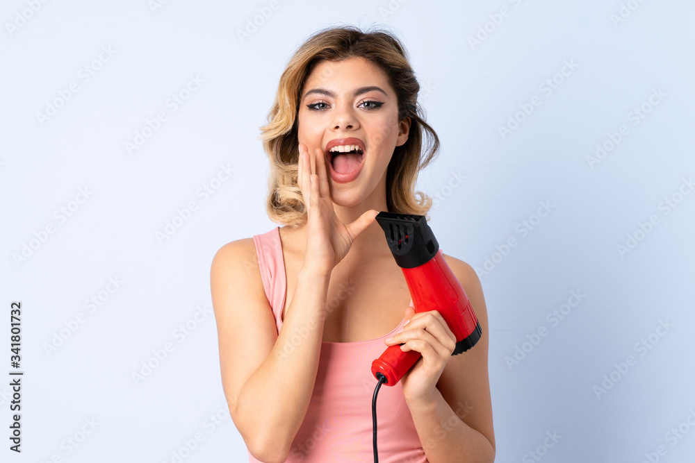Russian girl holding a hairdryer isolated on blue background shouting with mouth wide open Russian girl holding a hairdryer isolated on blue background shouting with mouth wide open