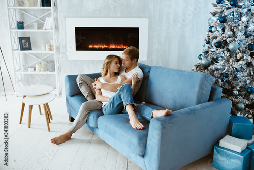 The guy and the girl hug and kiss on the blue couch. Christmas tree decorated with toys. Electronic fireplace. Bright interior. Blonde in a white T-shirt and jeans.