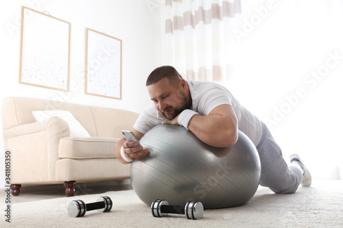 Foto Lazy overweight man using smartphone while lying on exercise ball at home