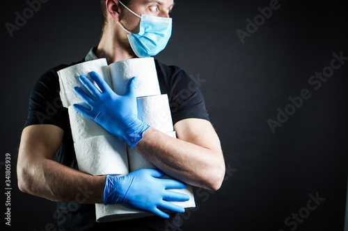 A young man dressed in a black T-shirt with blue rubber gloves and a protective mask on his face holds a pile of toilet paper in both hands. He's making a supply and prepares for a COVID-19 crisis.