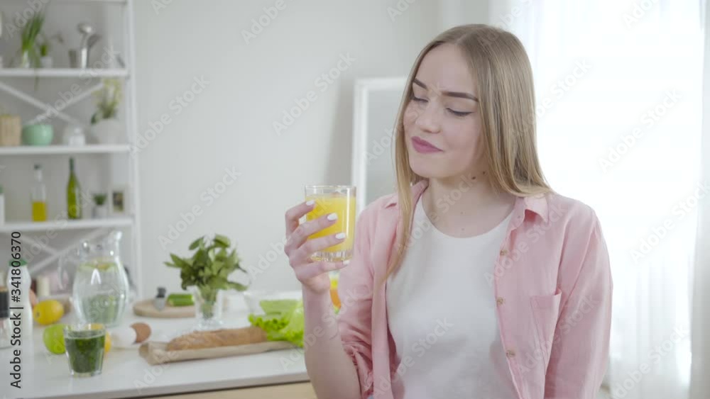 Blond young Caucasian woman drinking orange juice and smiling at camera. Portrait of fit pretty girl enjoying tasty organic drink indoors. Healthy eating, joy, lifestyle, happiness, beauty.