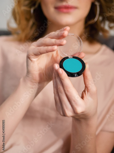 Woman's beauty blogger hand holding professional hi-end cosmetic trendy teal color eyeshadows box with blurred body background, warm pastel cozy tones and copyspace