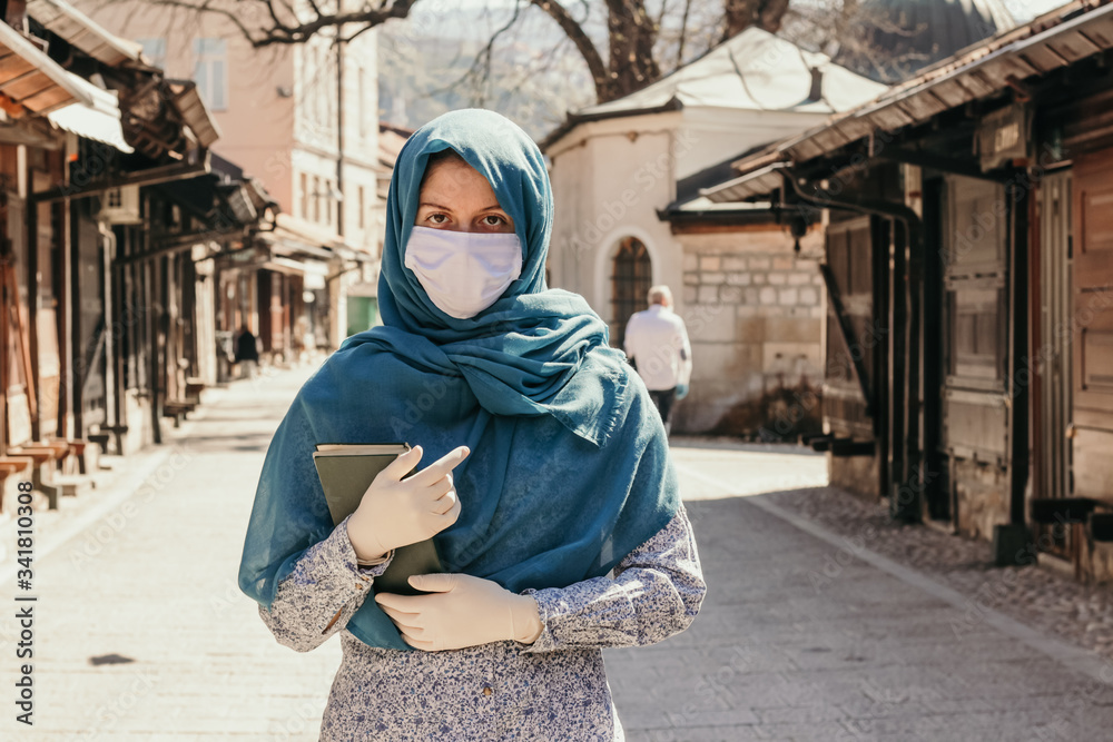 Young Muslim woman standing in front of a closed mosque during Ramadan due to the Coronavirus global pandemic. Ramadan 2020.  Sad woman holding Qur'an. Islamic country culture.