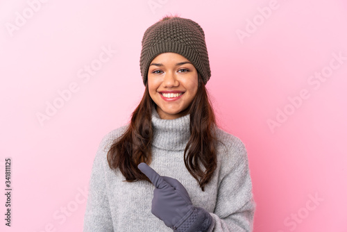 Young Colombian girl with winter hat over isolated pink wall pointing finger to the side