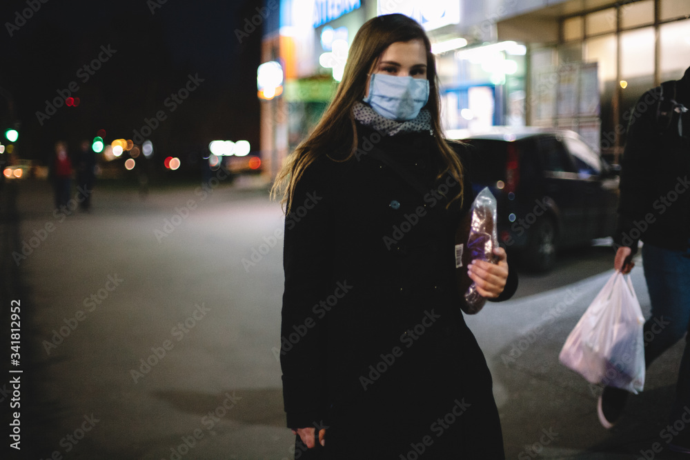 Young woman wearing face medical mask carrying bread while walking on city street at night Young woman wearing face medical mask carrying bread while walking on city street at night