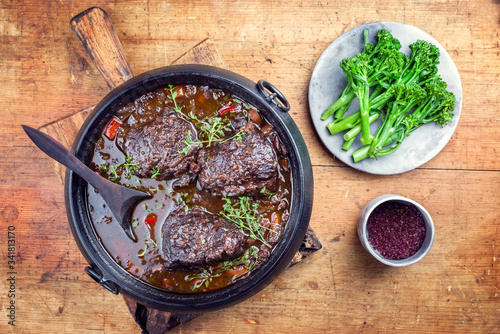 Traditional German braised beef cheeks in brown red wine sauce with carrots and broccoli offered as top view in a modern design stewpot on an old rustic board