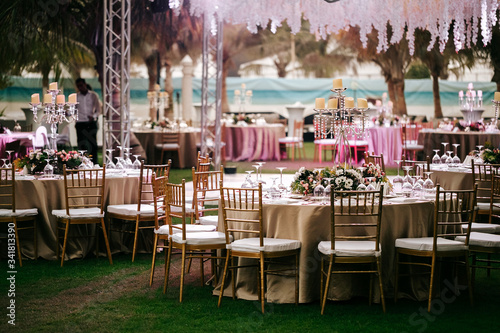 International Wedding outdoor celebration EVENING party under palm trees. Served tables on green area in hotel. Landyard. Beige and pink colors. Close-up and wide angle. © Maksim Denisenko