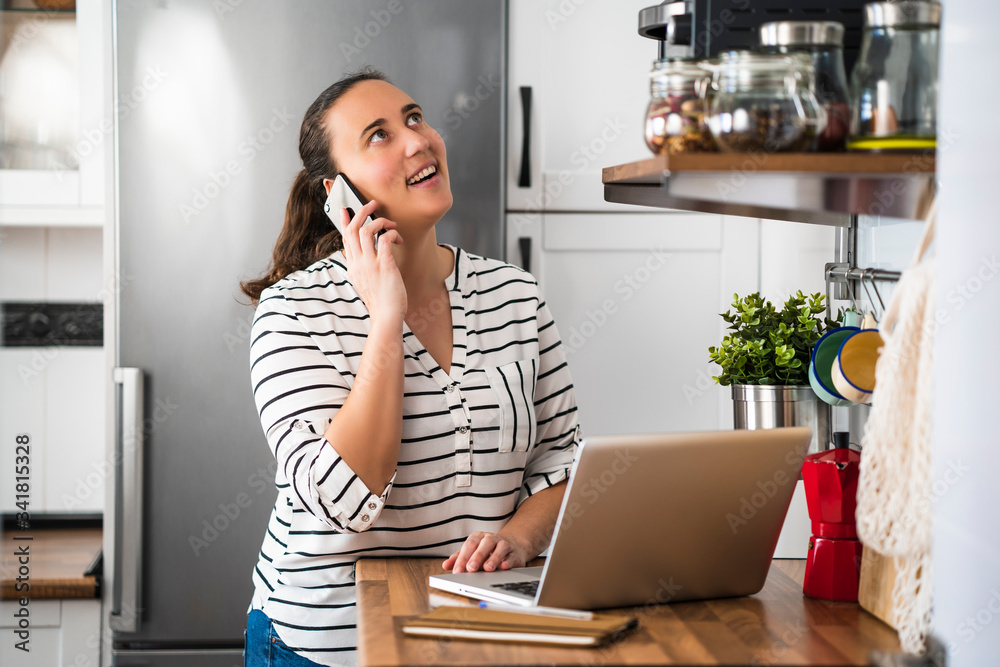 Happy young woman talking on her smartphone and using laptop in her apartment kitchen. Working at home and staying at home. Happy young woman talking on her smartphone and using laptop in her apartment kitchen. Working at home and staying at home.