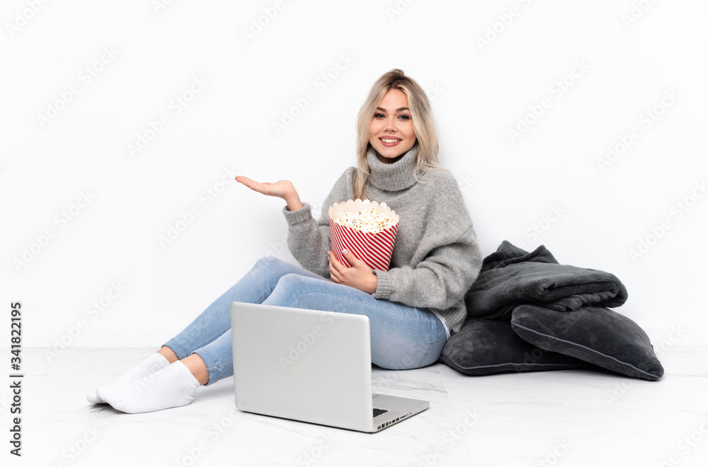 Teenager blonde girl eating popcorn while watching a movie on the laptop holding copyspace imaginary on the palm to insert an ad Teenager blonde girl eating popcorn while watching a movie on the laptop holding copyspace imaginary on the palm to insert an ad