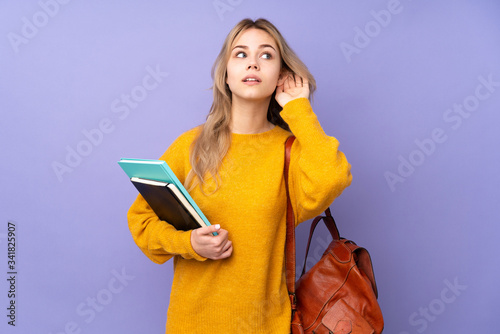 Teenager Russian student girl isolated on purple background listening to something by putting hand on the ear