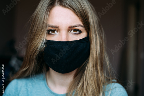 A young woman in a black medical mask with blond hair and dark eyes and a turquoise t-shirt is looking at the camera. portrait photo. quarantine. stay home concept. epidemic. virus. coronavirus.
