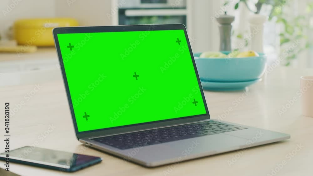 Close Up Green Screen Mock Up on a Laptop Computer. Device is Working on a Kitchen Table in a Modern Home. Smartphone Lies Next to the Laptop. Sunny Modern Kitchen with Healthy Lifestyle Vibes. Close Up Green Screen Mock Up on a Laptop Computer. Device is Working on a Kitchen Table in a Modern Home. Smartphone Lies Next to the Laptop. Sunny Modern Kitchen with Healthy Lifestyle Vibes.