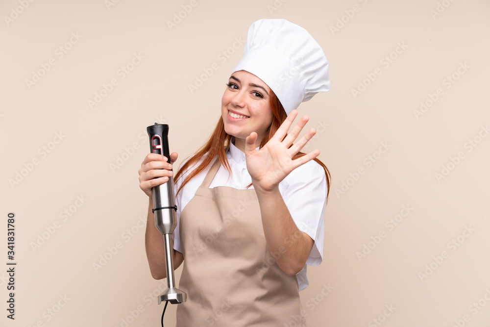 Redhead teenager girl using hand blender over isolated background saluting with hand with happy expression Redhead teenager girl using hand blender over isolated background saluting with hand with happy expression