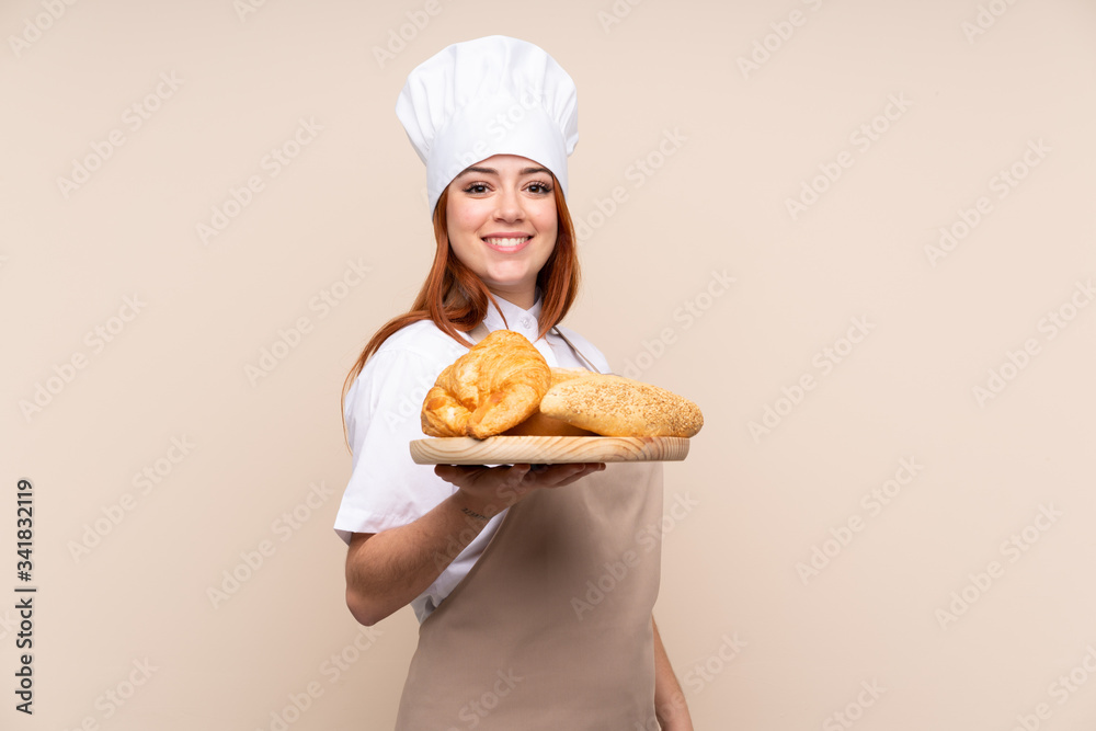 Redhead teenager girl in chef uniform. Female baker holding a table with several breads smiling a lot Redhead teenager girl in chef uniform. Female baker holding a table with several breads smiling a lot