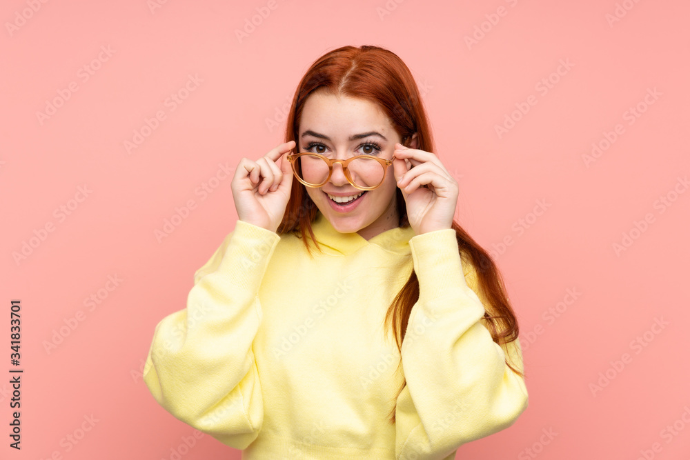 Redhead teenager girl over isolated pink background with glasses and surprised