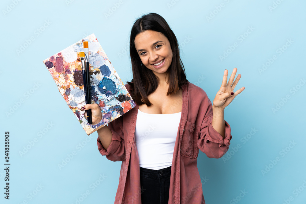 Young artist woman holding a palette over isolated blue background happy and counting four with fingers. Young artist woman holding a palette over isolated blue background happy and counting four with fingers.