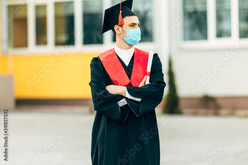 A male graduate student wears a protective mask against coronavirus, in a black graduation dress, with a diploma in his hands. quarantine, coronavirus