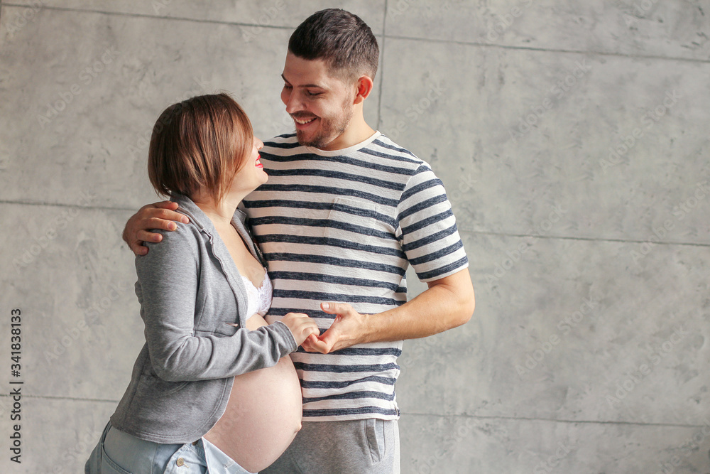 Beautiful smiling couple hugging in the Studio on a gray background. A pregnant girl in a gray sweatshirt with an open stomach. The guy in the striped tee shirt.