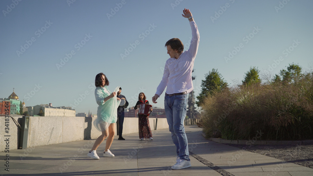 Fototapeta premium A young couple dancing to live music on the street. Musicians play, and a young couple dancing on the promenade. Music street performers couple with girl violinist and man guitarist on blue sky