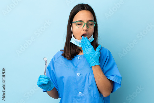 Young brunette mixed race dentist woman holding tools over isolated background yawning and covering wide open mouth with hand