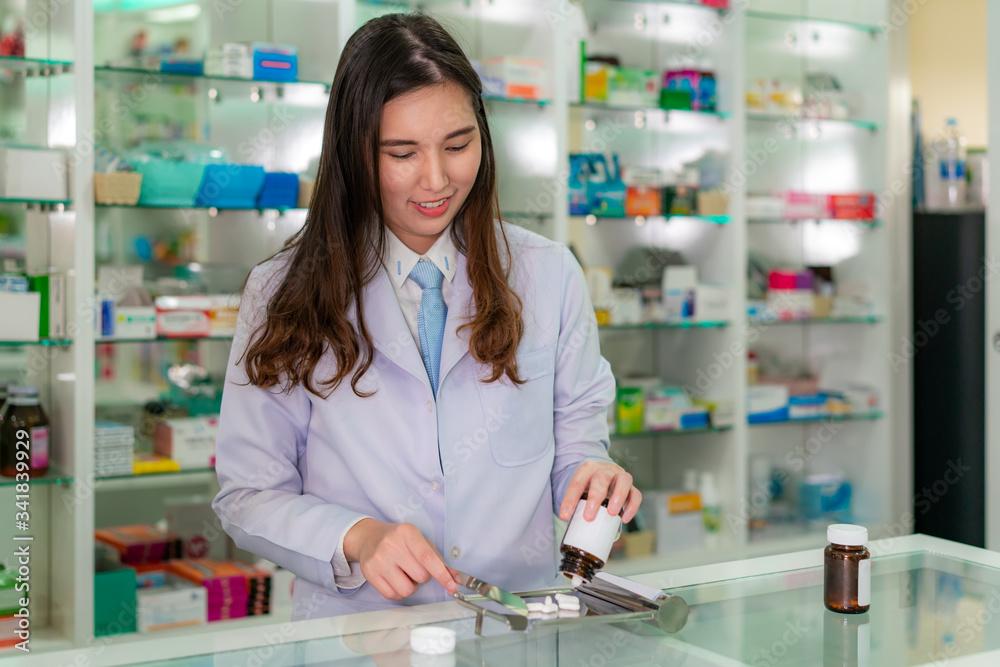 Fototapeta premium Azjatycka młoda farmaceutka wylewa i liczy kapsułki leków na nierdzewną tacę, przygotowuje opakowanie leku dla pacjenta w aptece. Koncepcja medycyny, farmacji, opieki zdrowotnej i ludzi.
