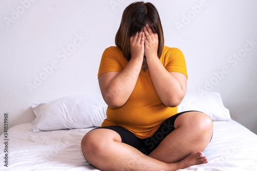 Overweight young woman sitting on white bed while holding hands cover on her face at home. Upset female suffering from extra weight in the bedroom. Obesity unhealthily concept. photo