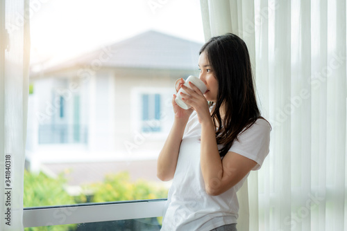 Beautiful Asian woman wake up in her bed fully rested and open the curtains on the windowsill and looking out the window with cup of coffee in the morning in bedroom at home..