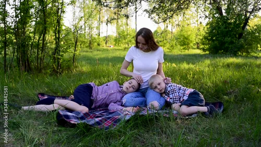 Two sons put his head on mother's lap and mom stroked their heads. Mother with two children sitting outdoors on picnic blanket