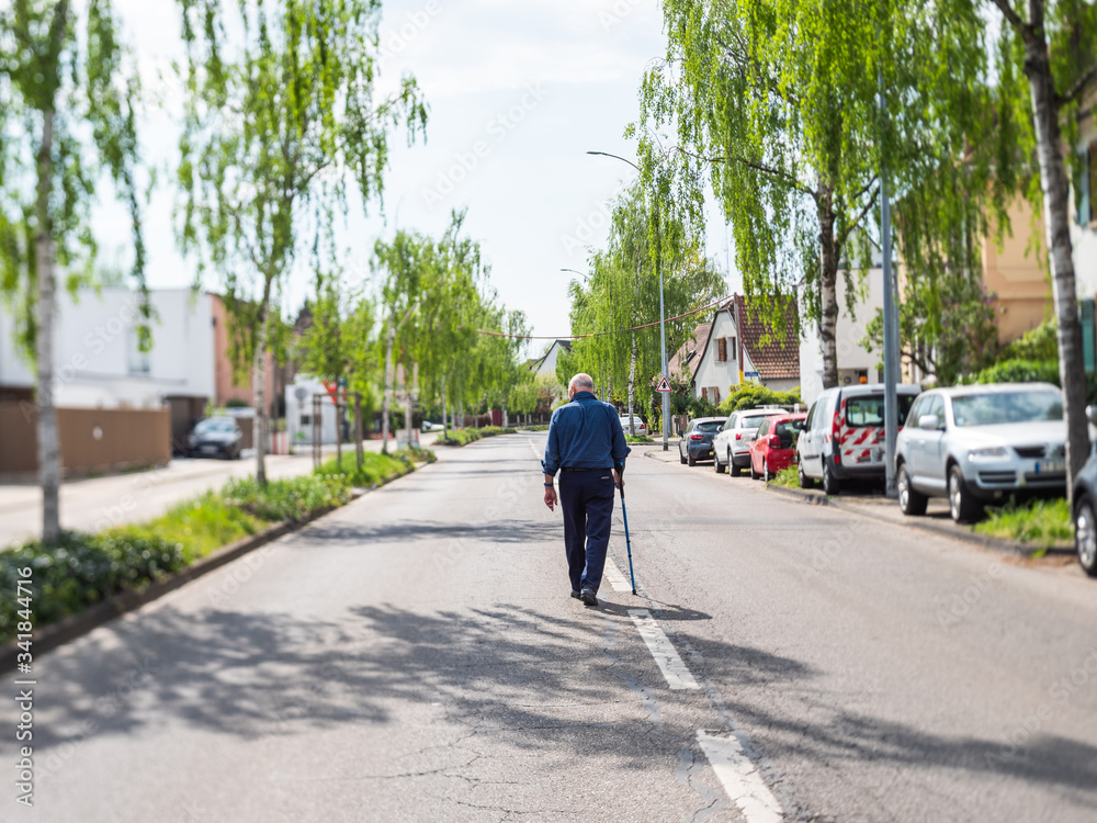Perspective view over a birch alley street with lonely senior man using a cane walking alone in the middle of the street during coronavirus covid-19 lockdown in France - tilt-shift lens