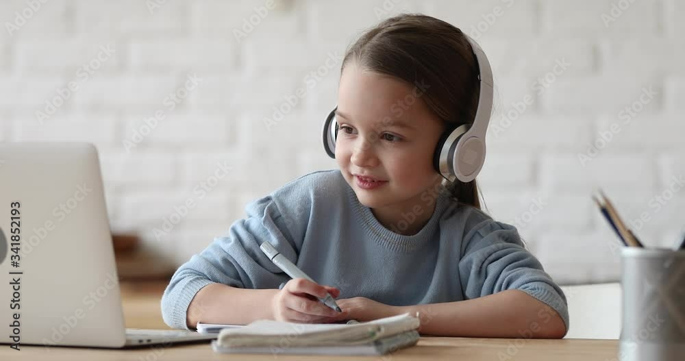 Happy small primary school girl elearning online using computer app. Child pupil wearing headphones video calling remote tutor or watching video lesson class on laptop. Kid distance study at home.