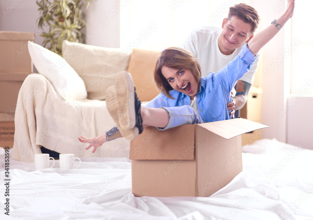 Couple having fun laughing moving into new home, young woman riding sitting in cardboard box while man pushing it