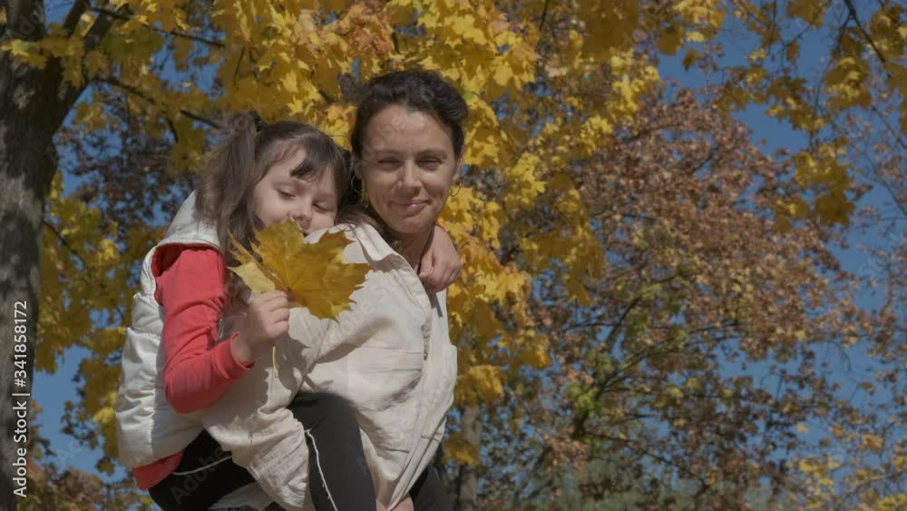 Mother hold child in yellow park. A cute little girl with autumn leaves sits on her mother's back. Mother hold child in yellow park. A cute little girl with autumn leaves sits on her mother's back.