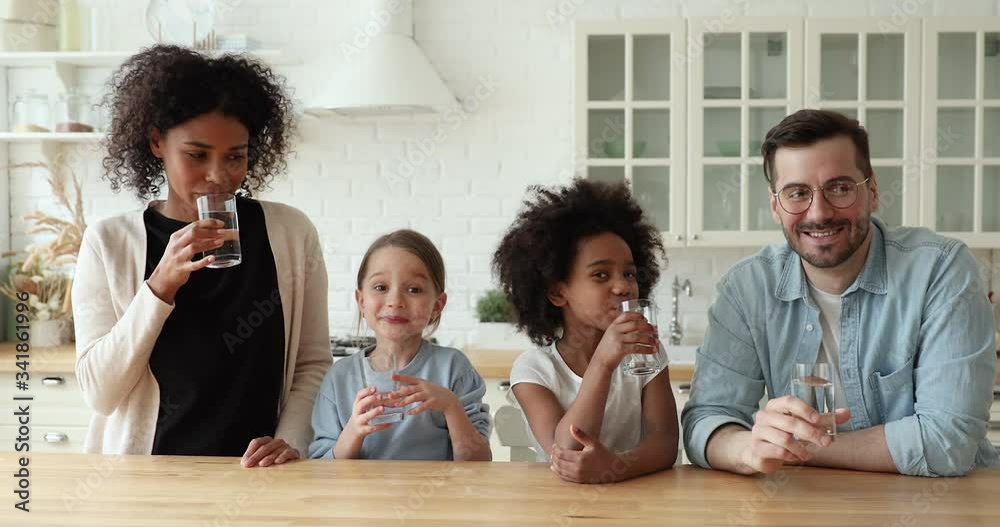 Happy healthy mixed race family young interracial parents and kids daughters drinking clean fresh pure mineral water showing thumbs up recommend good quality looking at camera in kitchen. Portrait.