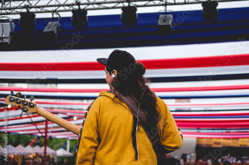 Bassist from back with bass, black cap and yellow sweatshirt playing live under red, blue and white decoration