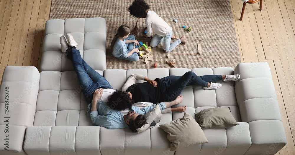 Mixed ethnicity parents relaxing on comfortable couch while cute diverse kids daughters playing on floor at home. Interracial family with two children spending time together in living room. Top view