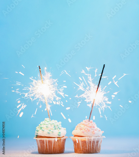 Two cupcakes decorated with colorful sprinkles and sparklers. Festive cupcakes with blue and pink sprinkles on blue background with copy space top for text or design