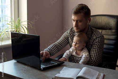 Real photo of young serious caucasian man work at home office on laptop with he baby daughter during the quarantine. Work at home, maternity leave for father
