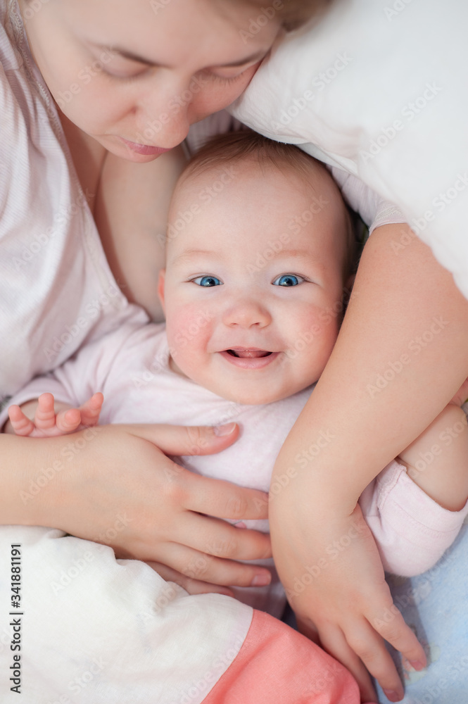 Happy mom and daughter lie on their backs. Happy smiling baby lying on bed at home. Beautiful smiling girl with blue eyes. Top view. Flat Lay Happy mom and daughter lie on their backs. Happy smiling baby lying on bed at home. Beautiful smiling girl with blue eyes. Top view. Flat Lay