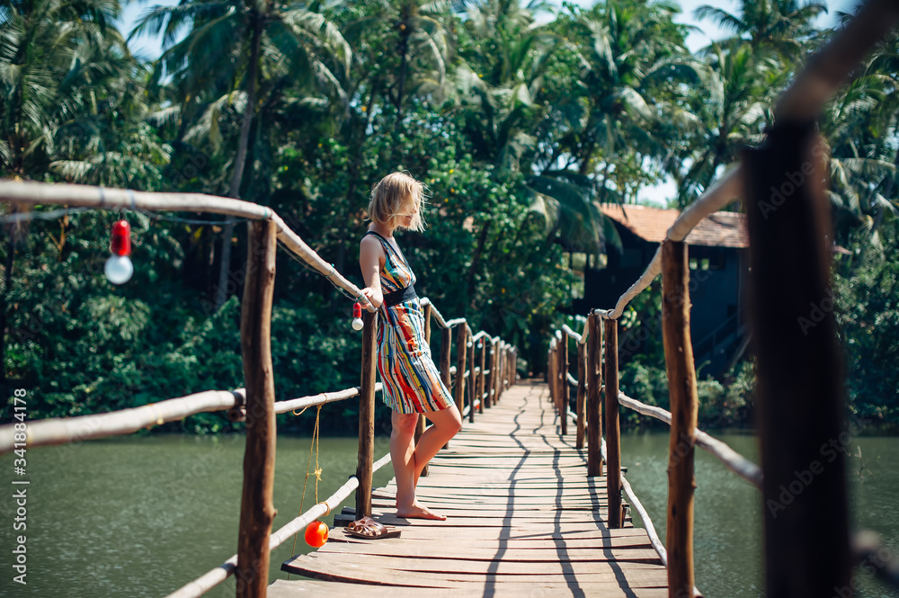 Fototapeta premium A female tourist in bright dress on sunny tropical day. Young beautiful girl stands on wooden bridge over small river against blurred palm jungle. Picturesque landscape with copy space.