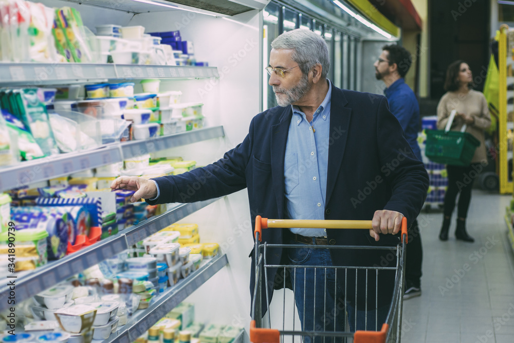 Mature man choosing dairy products in supermarket. Serious man with shopping trolley looking at shelves in refrigerator while buying goods in store. Shopping concept