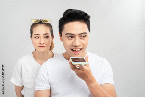 Portrait of a young couple using mobile phones while standing together over gray background, curious woman looking at mans phone