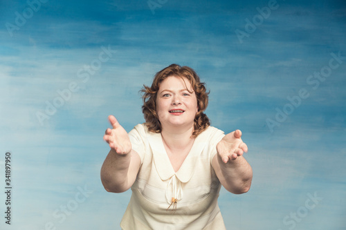 Redhead funny overweight young woman screams something, bulging her eyes and waving his arms on a blue background.