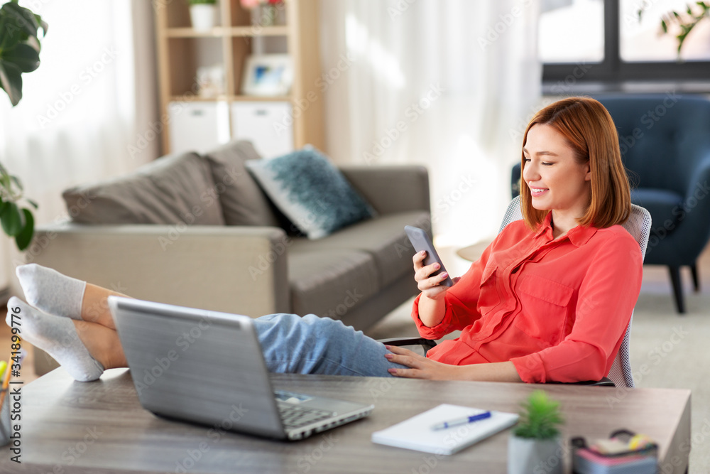 remote job, technology and people concept - happy smiling young woman with smartphone and laptop computer at home office resting feet on table