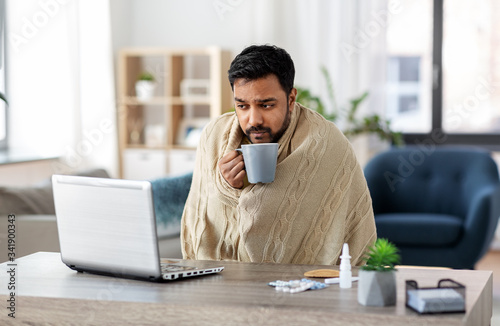 Fototapeta health, cold and people concept - sick young indian man in blanket with laptop c
