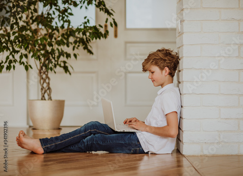 Young  blond boy in white shirt and jeans printing on the white laptop sitting on the floor at home