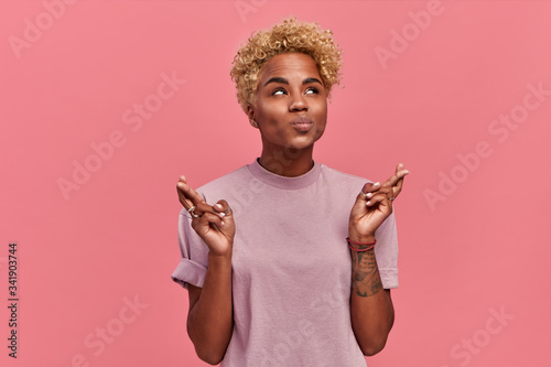 Konstfotografi Surprised young blonde dark-skinned woman crosses fingers, makes praying gesture, dressed in casual summer clothes, isolated over pink background